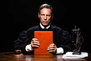 judge in judicial robe sitting at table and holding orange book isolated on black