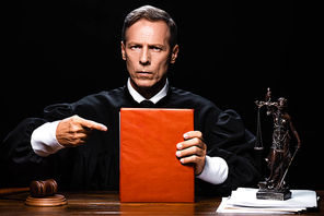 judge in judicial robe sitting at table and pointing with finger at orange book isolated on black