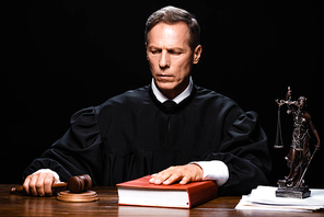 judge in judicial robe holding gavel and putting hand on book isolated on black