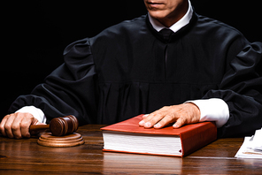 cropped view of judge in judicial robe sitting at table and holding gavel and book isolated on black