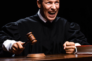 angry judge in judicial robe sitting at table and hitting with gavel isolated on black