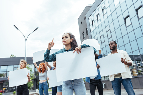 selective focus of attractive woman pointing with finger while holding blank placard near multicultural people