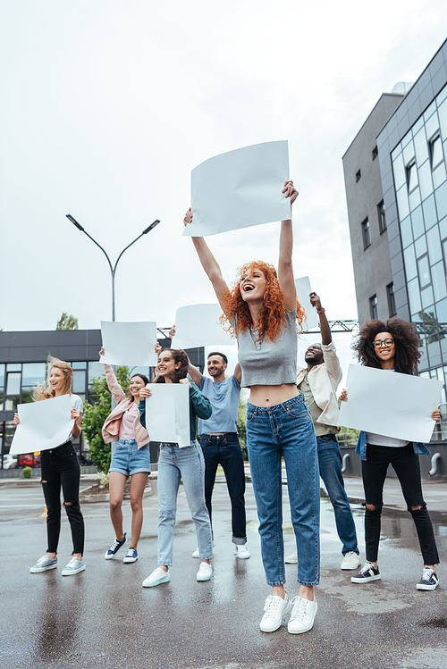 selective focus of happy redhead girl holding empty board near multicultural people