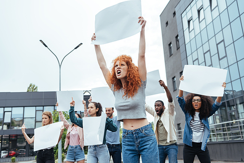 selective focus of angry redhead girl holding blank placard on meeting near multicultural people