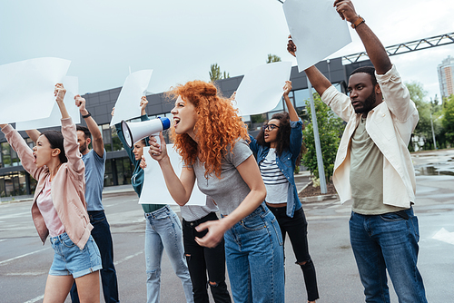 emotional redhead girl screaming in megaphone near multicultural people