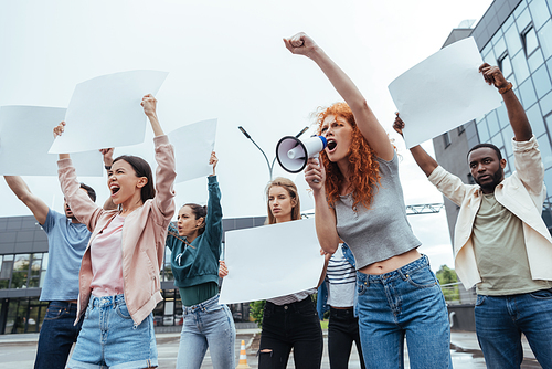 emotional redhead woman screaming in megaphone near multicultural people