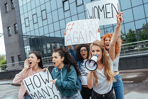 selective focus of emotional girl holding megaphone and gesturing near multicultural women holding placards on meeting