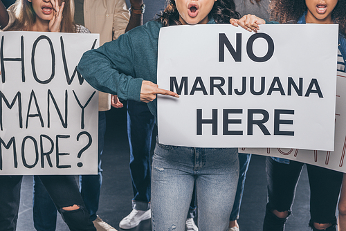 cropped view of emotional girl pointing with finger at placard with no marijuana here lettering near multicultural people on black