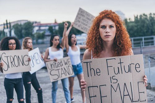 selective focus of redhead girl holding placard with future is female letters near multicultural girl outside