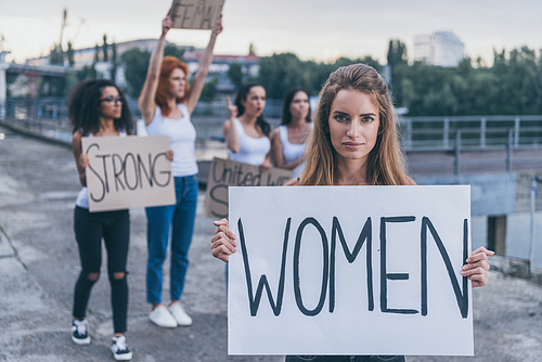 selective focus of woman holding board with women lettering near multicultural girls outside