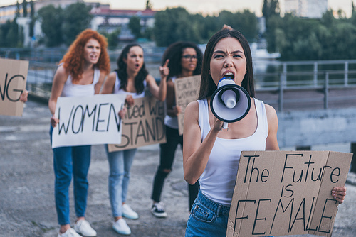 selective focus of emotional girl holding placard with the future is female letters and screaming in megaphone outside