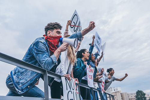 low angle view of emotional man with scarf on face gesturing and holding megaphone near multicultural people with placards