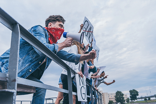 man with scarf on face gesturing and holding megaphone near multicultural people with placards