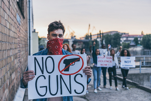selective focus of man with scarf on face holding placard with no guns lettering near multicultural people