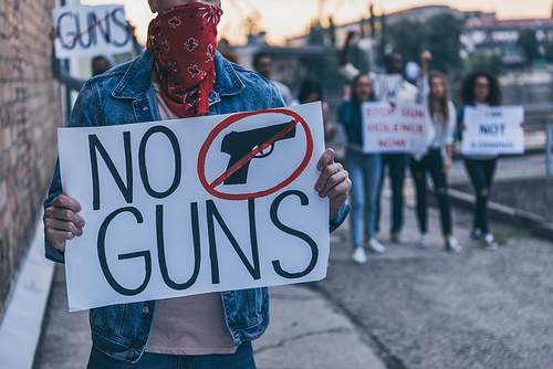 cropped view of man with scarf on face holding placard with no guns lettering near multicultural people