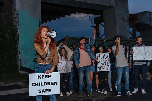 selective focus of emotional redhead girl holding placard with keep children safe lettering and screaming near multicultural people on meeting