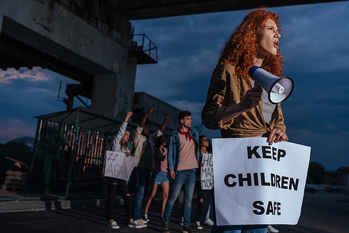 selective focus of emotional redhead girl holding placard with keep children safe lettering near multicultural people on meeting