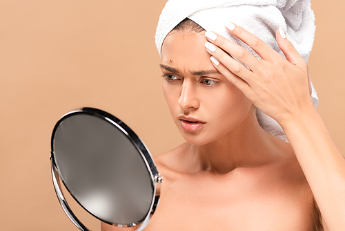 naked woman in towel looking at mirror and touching face with pimple isolated on beige