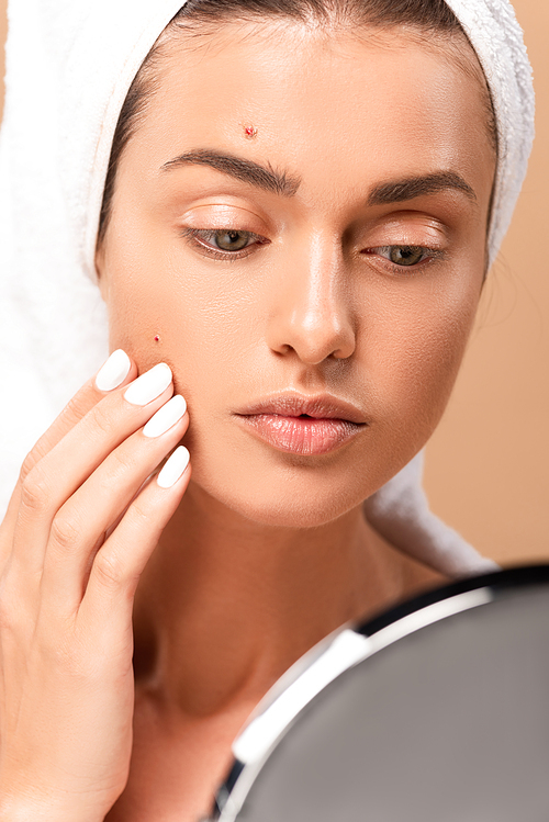 selective focus of girl in towel touching face with pimples and looking at mirror isolated on beige