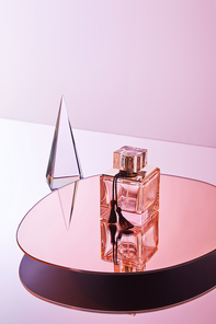 crystal transparent pyramid with reflection near perfume bottle on round mirror on pink background