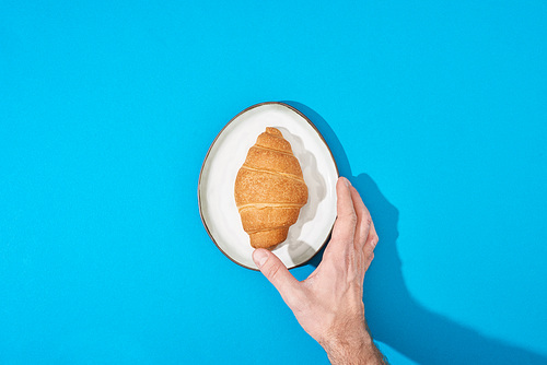 cropped view of man holding fresh croissant on plate on blue background