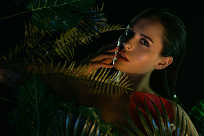 Attractive girl with flower touching face behind green leaves isolated on black