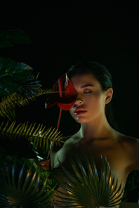 Woman with closed eyes covering face with flower near leaves isolated on black