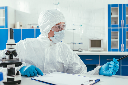 biochemist in hazmat suit sitting near microscope and clipboard in laboratory