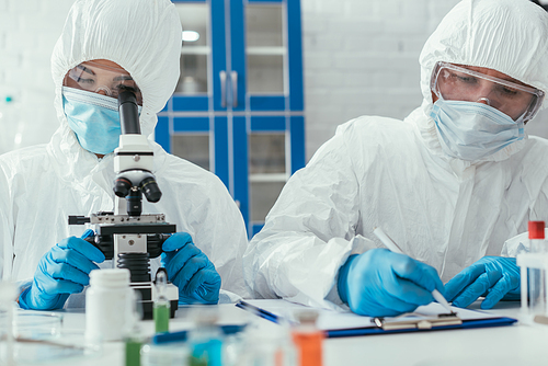 selective focus of biochemist looking through microscope near colleague writing down results on clipboard