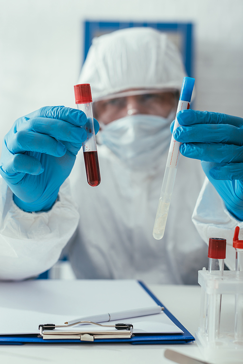 selective focus of biochemist in hazmat suit holding test tubes with blood sample and liquid