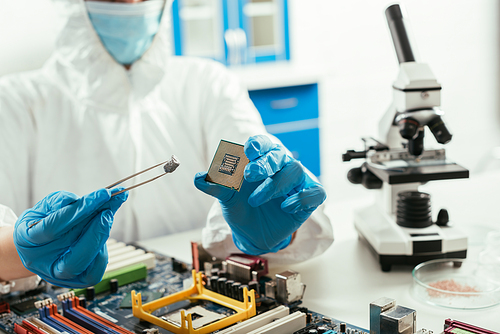 cropped view of engineer holding microchip and small stone near microscope and computer motherboard