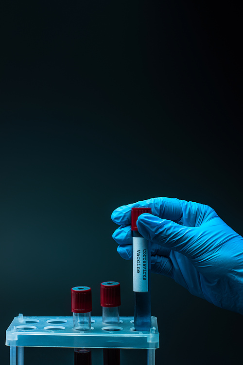 Cropped view of male hand with coronavirus vaccine and sample tubes with blood in test tube rack on dark background