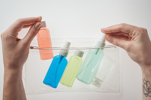 cropped view of woman putting bottles with liquids to cosmetic bag on white background