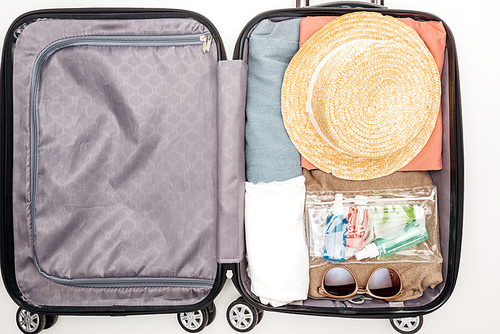 top view of travel bag with towel, cosmetic bag with bottles, clothes, sunglasses and hat on white background