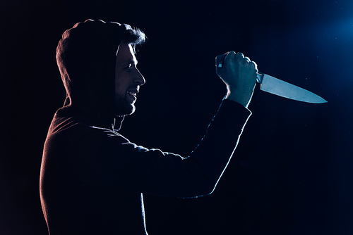 side view of smiling criminal in casual clothes holding knife on black