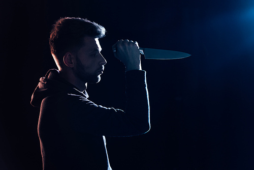 side view of pensive criminal in casual clothes holding knife on black