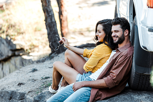 cheerful bearded man and attractive girl sitting near car and trees