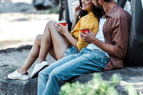 cropped view of bearded man and happy girl sitting near car and holding cups