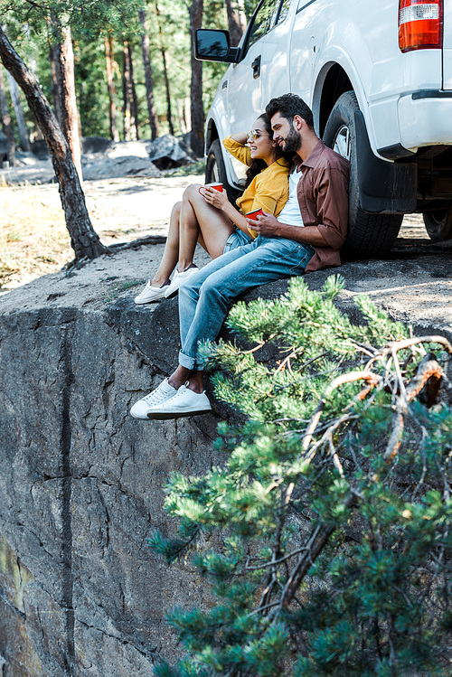 selective focus of bearded man and attractive girl sitting near car and holding cups in woods