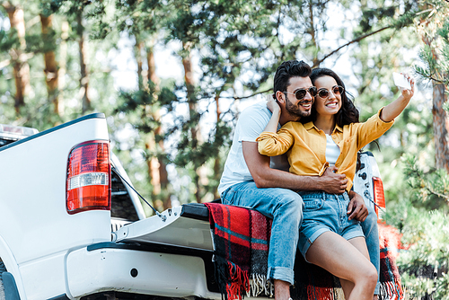 cheerful young woman talking selfie with bearded man near car and trees