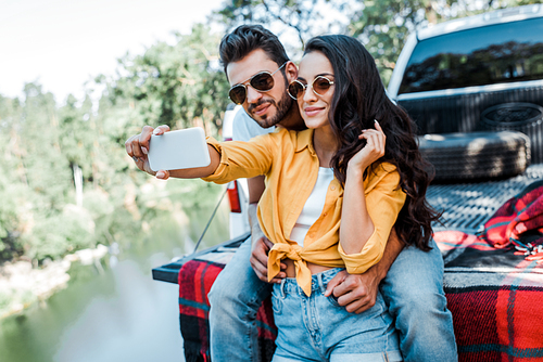 cheerful girl in sunglasses talking selfie with bearded man near car and lake