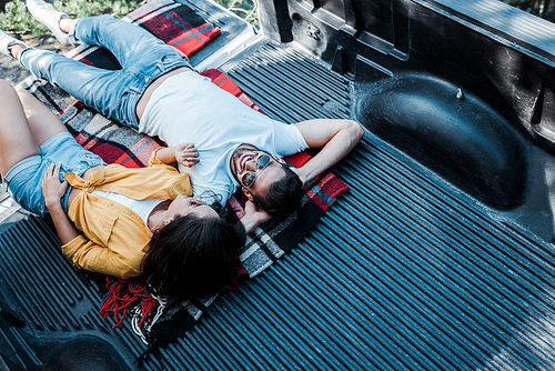 overhead view of woman in sunglasses lying with happy man in plaid blanket in car trunk