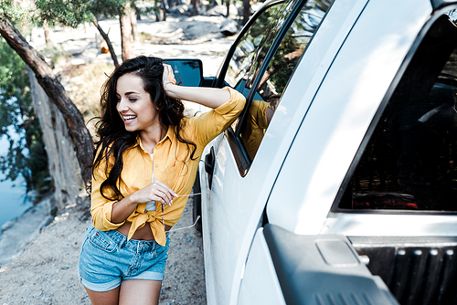 selective focus of happy woman smiling while standing near car in woods