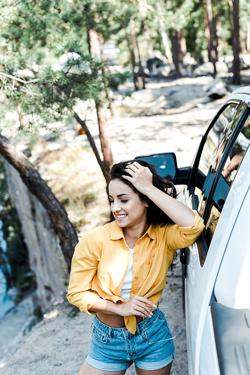 selective focus of attractive woman smiling while standing near car in woods