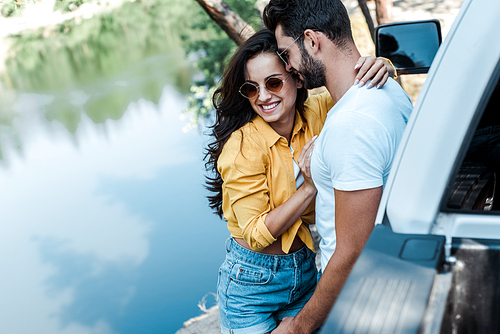 selective focus of happy girl hugging with boyfriend near car and river