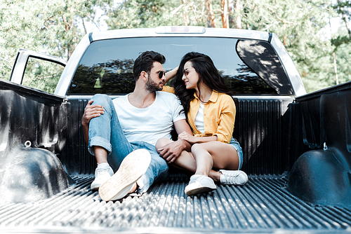 handsome man in sunglasses sitting in car trunk and looking at girl