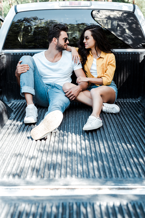 cheerful man in sunglasses sitting in car trunk and looking at girl
