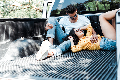 bearded man in sunglasses sitting in car trunk and looking at girl