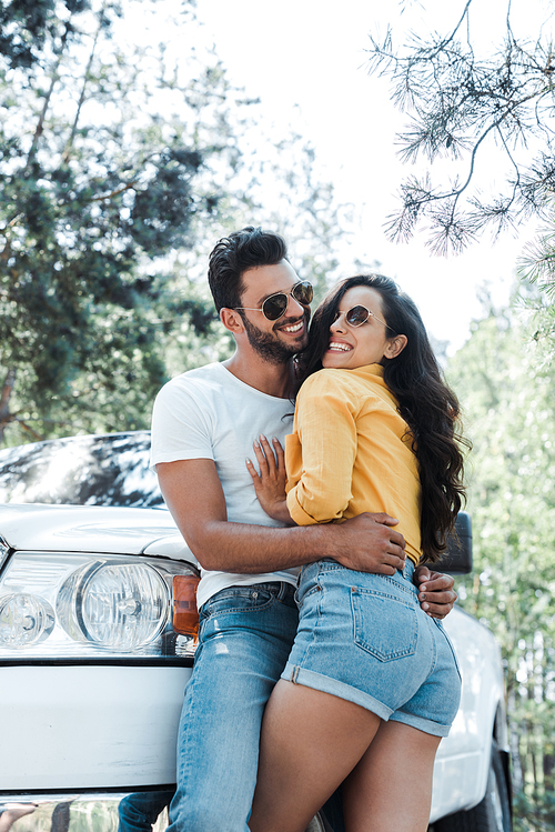 low angle view of man standing and hugging happy woman near auto in woods