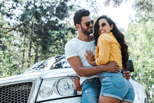 low angle view of man standing and hugging happy girl near auto in woods